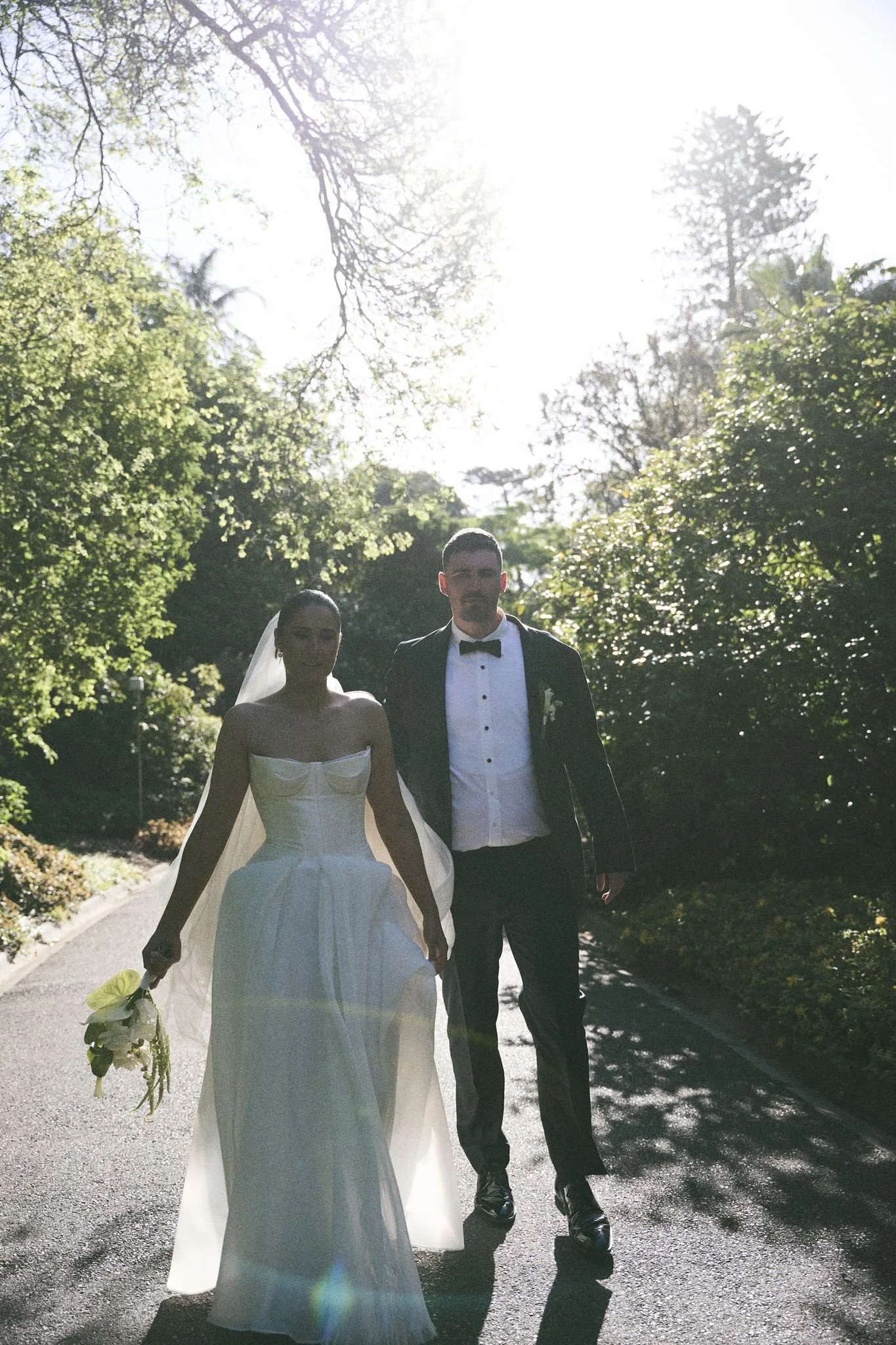 A bride and groom walking outdoors on a sunny day, surrounded by trees and greenery, holding hands.