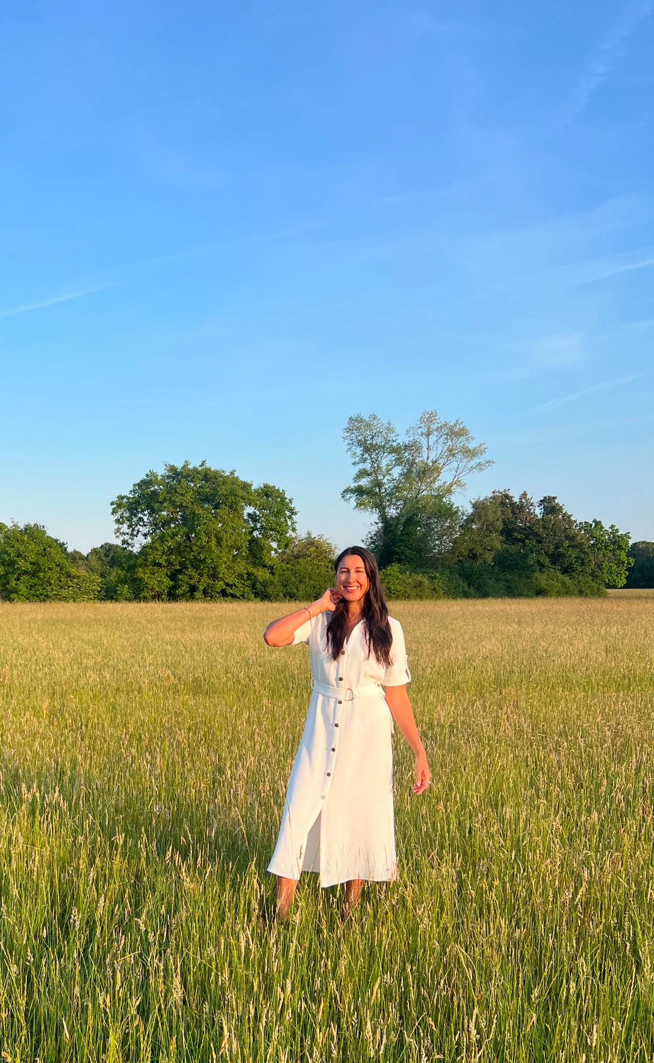 A woman in a white dress standing in a grassy field under a clear blue sky.