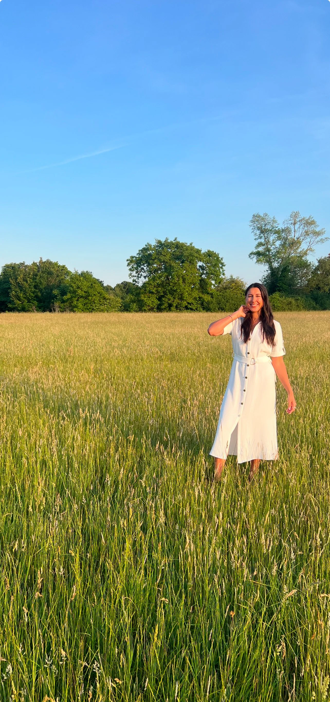 A woman in a white dress standing in a grassy field under a clear blue sky.