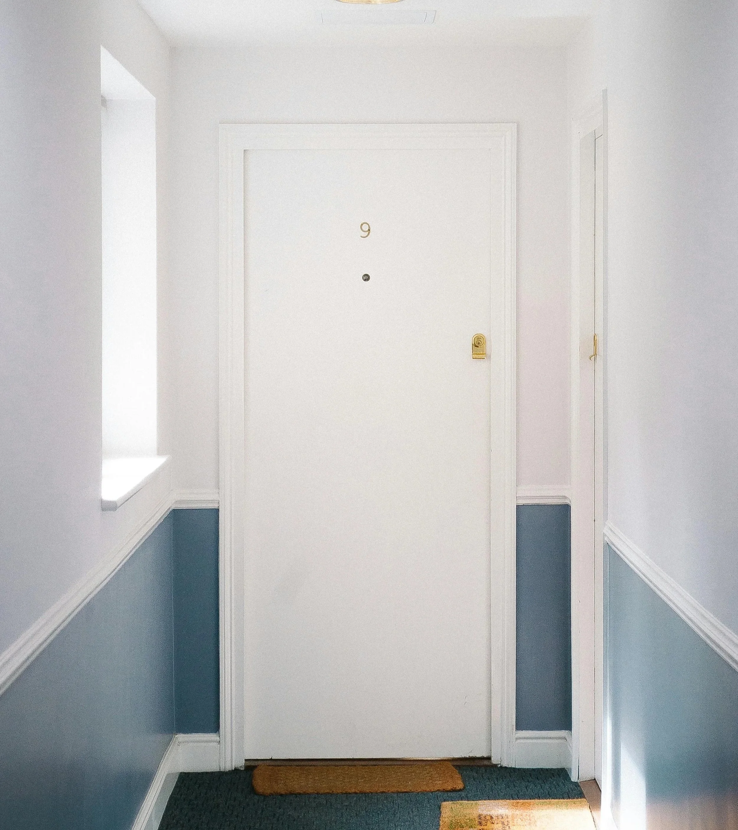 White apartment door with a gold number 9, peephole, and deadbolt lock, flanked by blue wainscoting and adjacent to a window and another door.