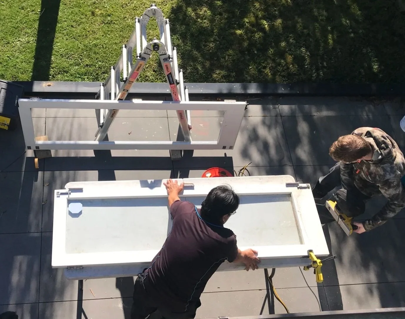 Two people working on a construction project outside, installing or repairing a white window frame on a black flat roof, with tools and a ladder nearby.
