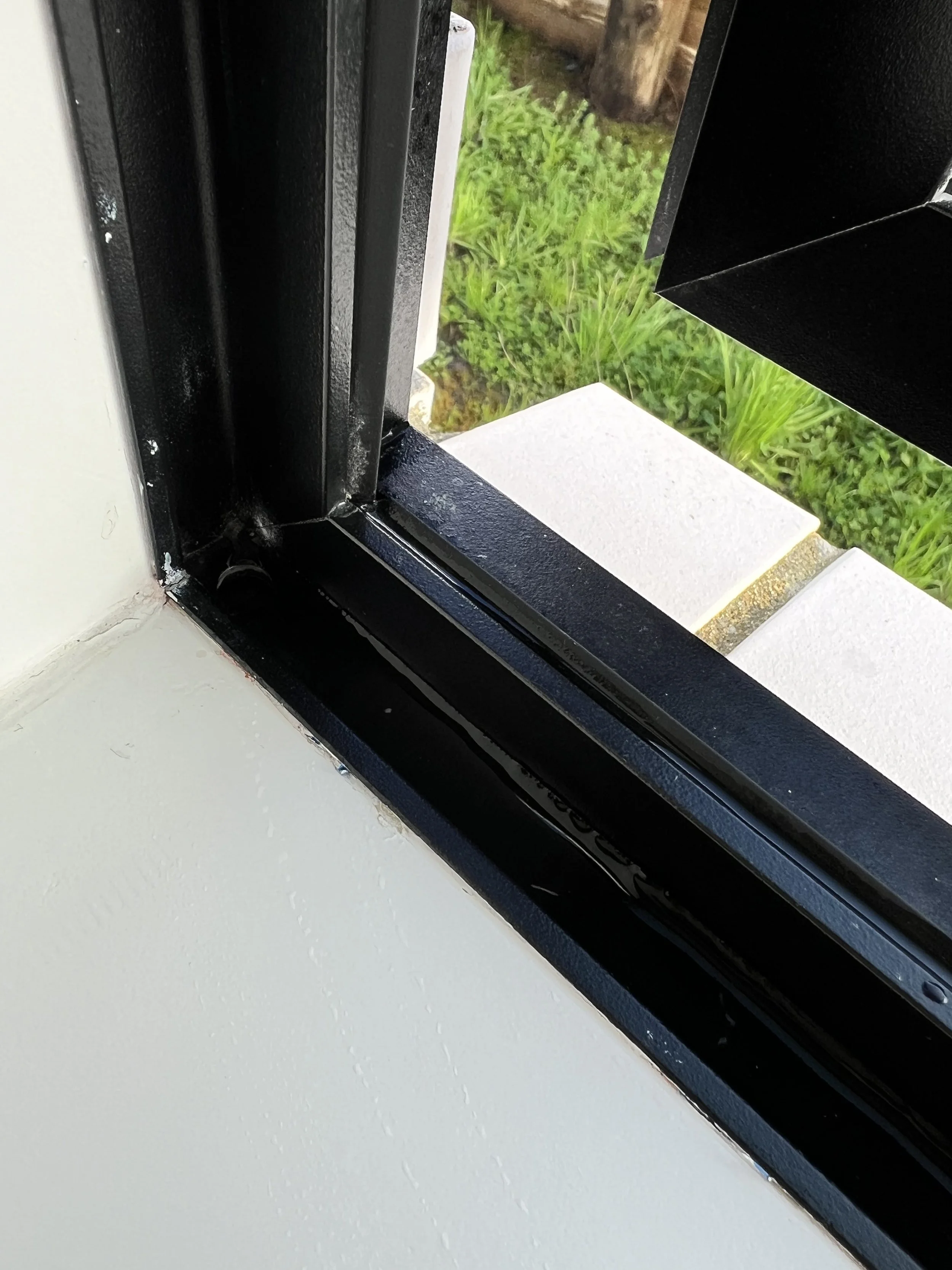 Close-up of a black window frame and sliding glass door, with grass and concrete sidewalk visible outside.