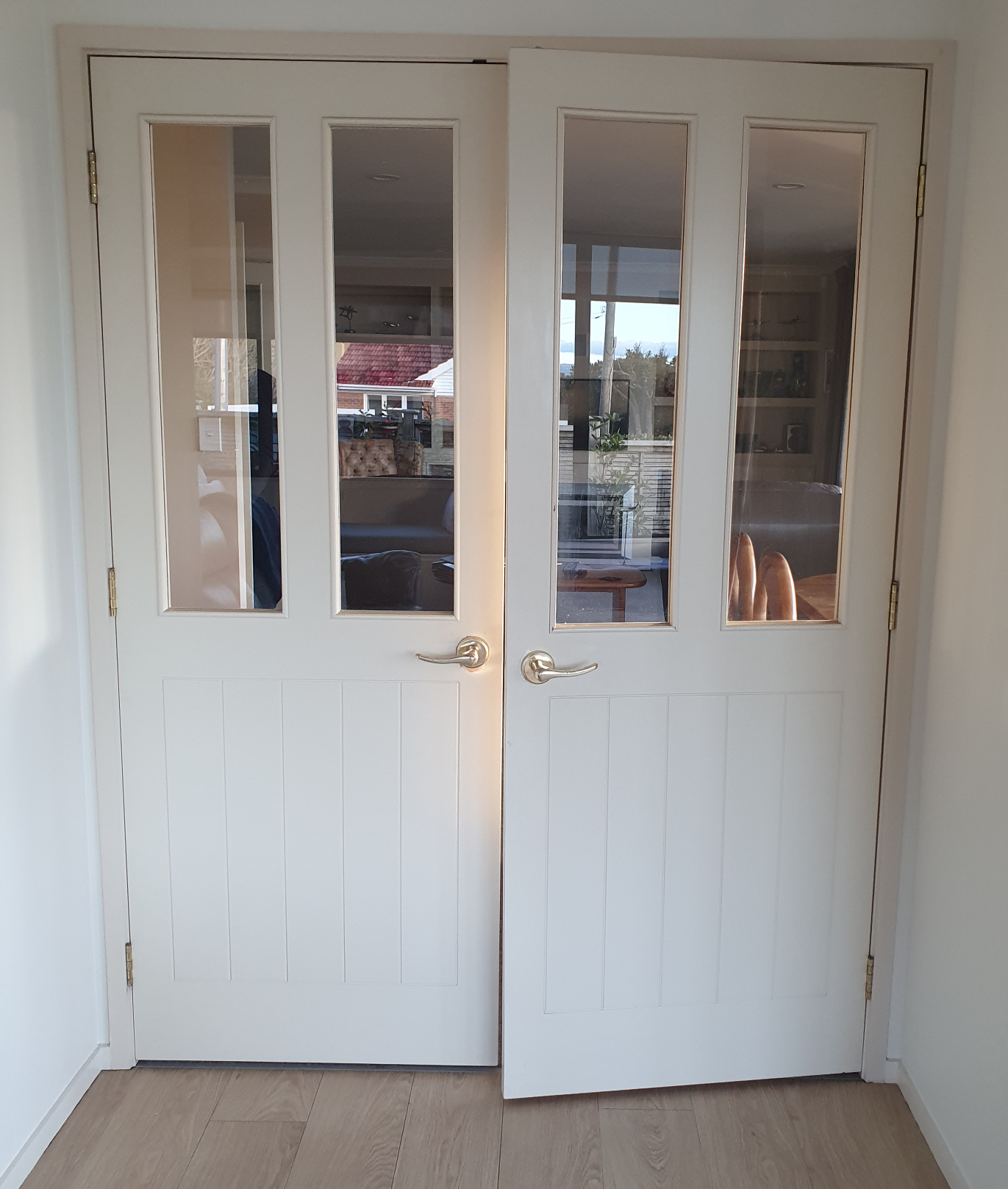 A set of white double doors with four glass panes in each door, brass handles, and brass hinges, leading to a room with visible furniture and windows.