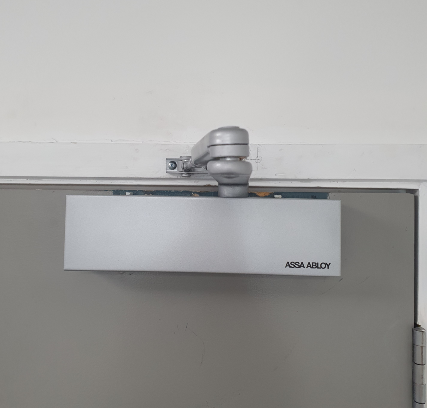 Fire door closer mounted above a gray door with the brand name 'ASSA ABLOY' printed on it.