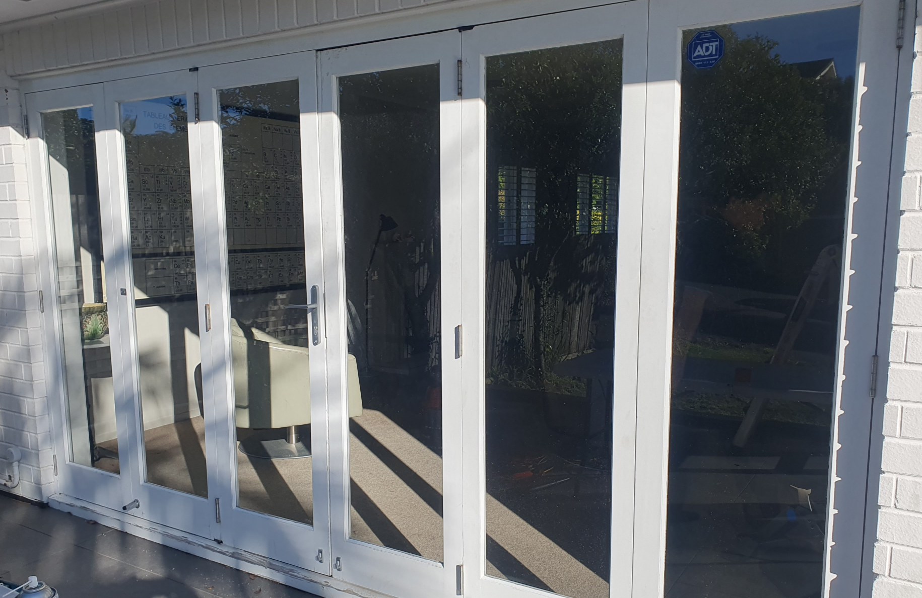 Outside view of a white-framed glass door with five panels, leading to an indoor space, with a white brick wall on the right. Reflections on the glass show greenery, a wooden fence, windows with blinds, and interior furniture, including a chair and exercise equipment.