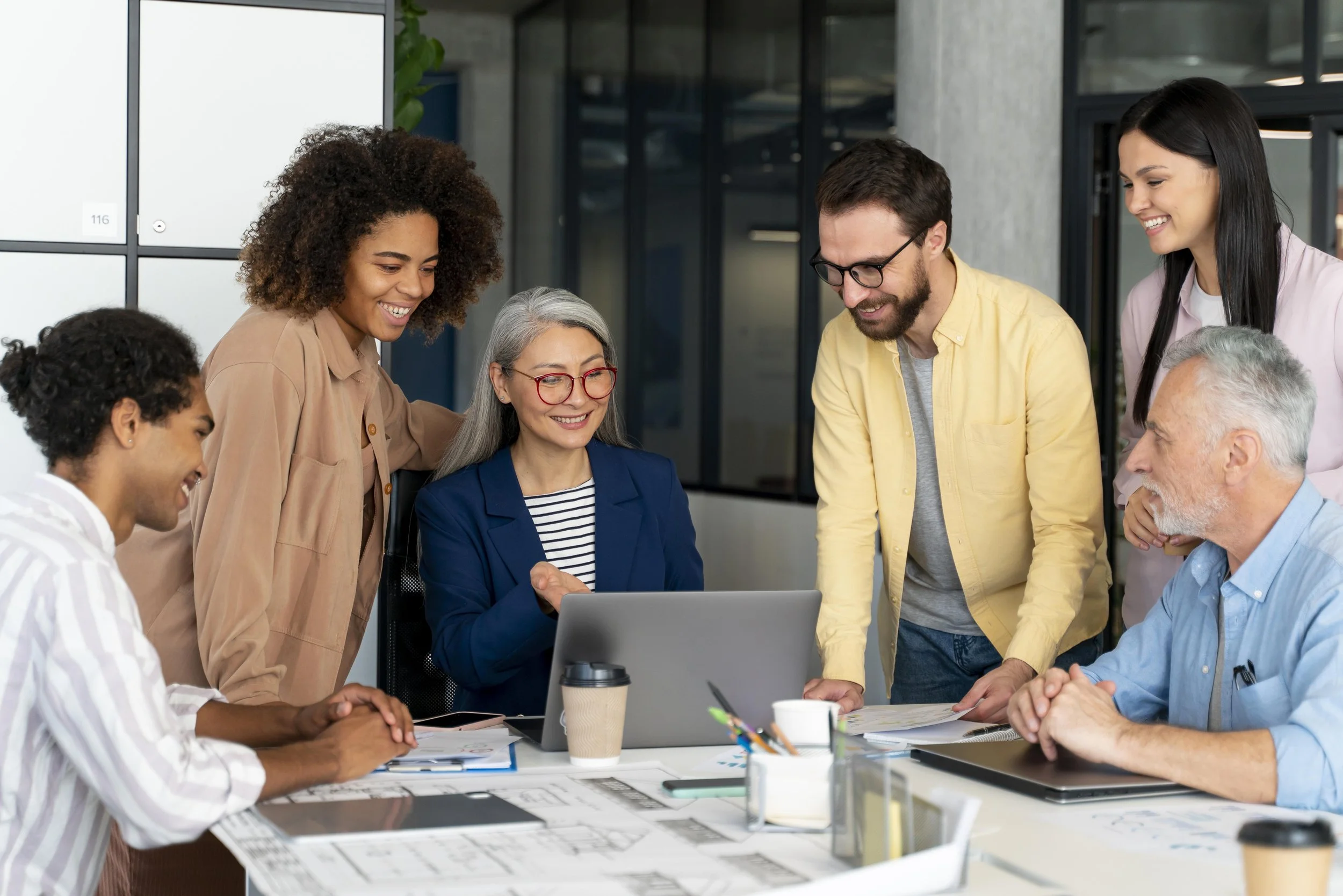A diverse group of people gathered around a laptop in a modern office, smiling and interacting during a meeting or discussion.