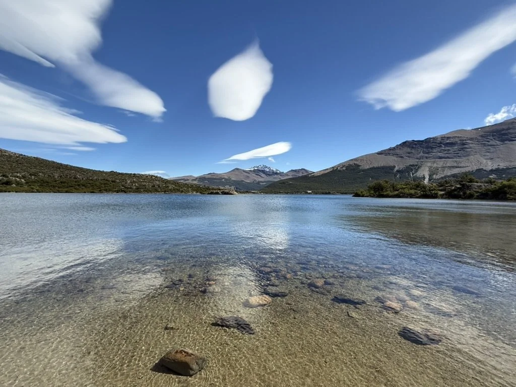 Clear shallow water at Laguna Capri beneath mountains and distinctive lenticular clouds in Patagonia.