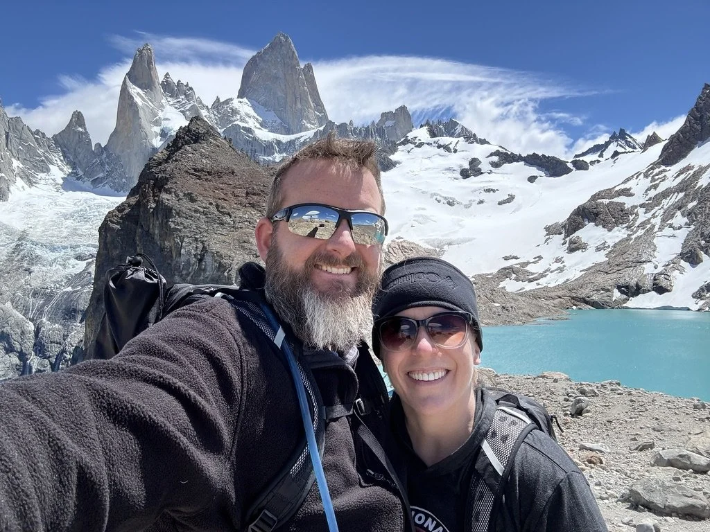 Couple smiling together with turquoise Laguna de los Tres and the snow-covered peaks of the Fitz Roy massif behind them.