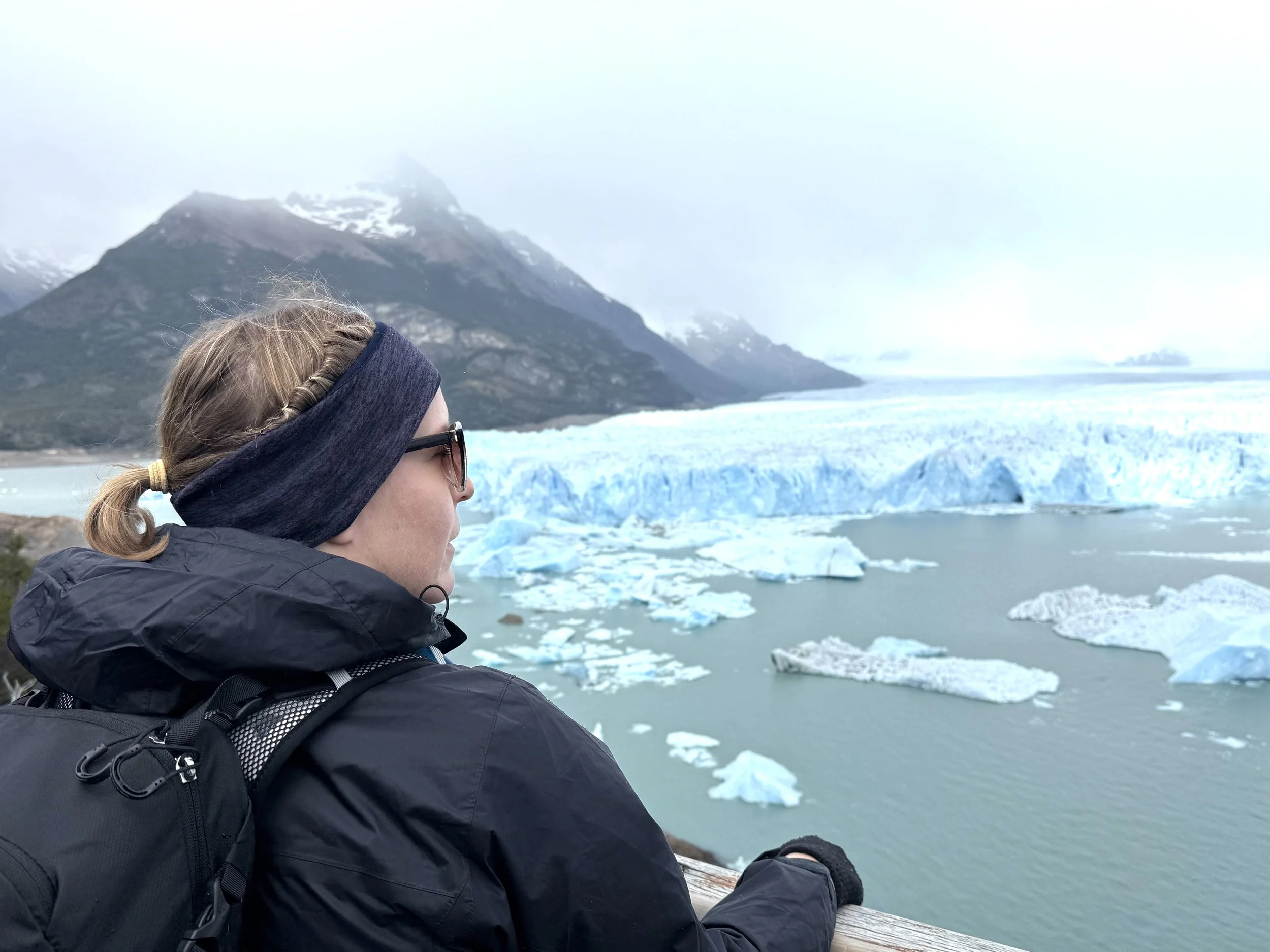 Person standing at a viewpoint overlooking the Perito Moreno Glacier, with blue ice and floating icebergs visible on Lago Argentino in Patagonia, Argentina.