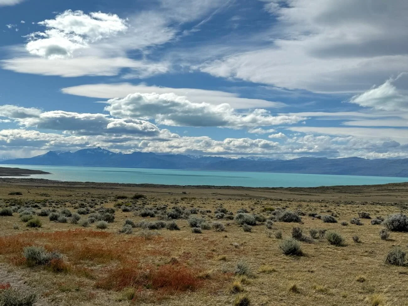 Wide view of Lago Argentino with turquoise water, Patagonian steppe in the foreground, and distant mountains under a cloudy sky in front of the Glaciarium Patagonia Ice Museum in El Calafate, Argentina.