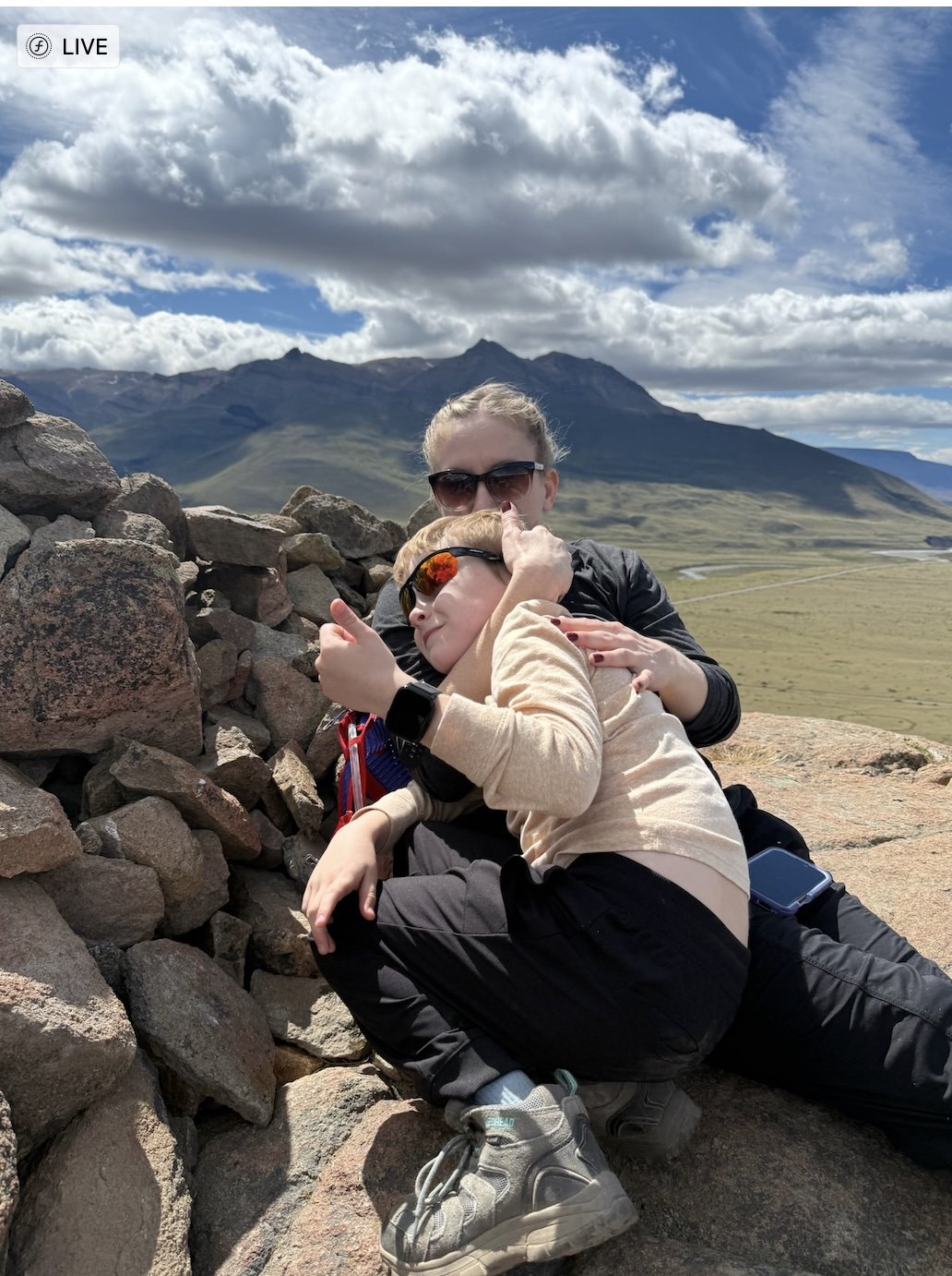 An adult and child resting together on rocks at the end of a hike near El Chaltén, Patagonia, with open steppe and mountains in the background.