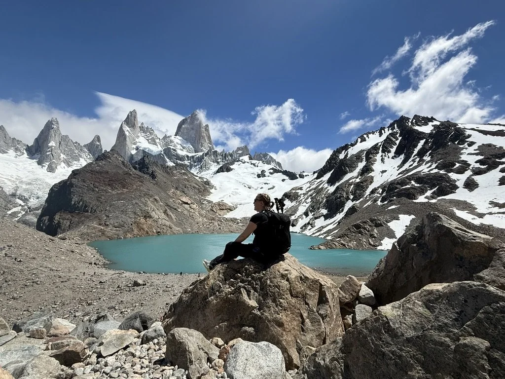 Hiker sitting on a boulder above the turquoise water of Laguna de los Tres with Mount Fitz Roy and snow-covered peaks behind her.