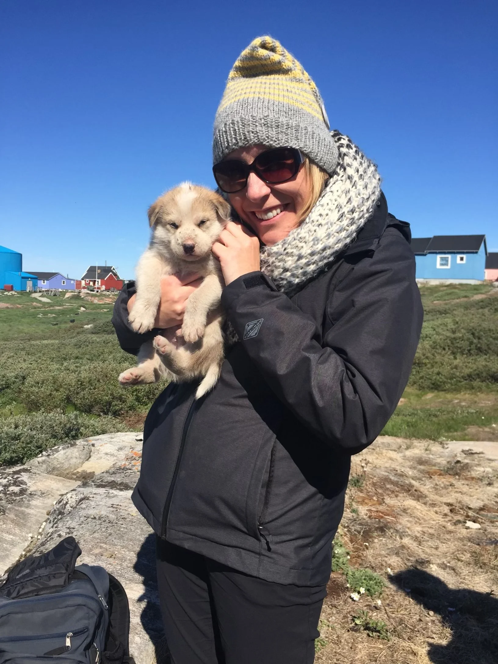 Traveler holding a sled dog puppy outdoors in Ilulissat, Greenland, with colorful houses in the background.