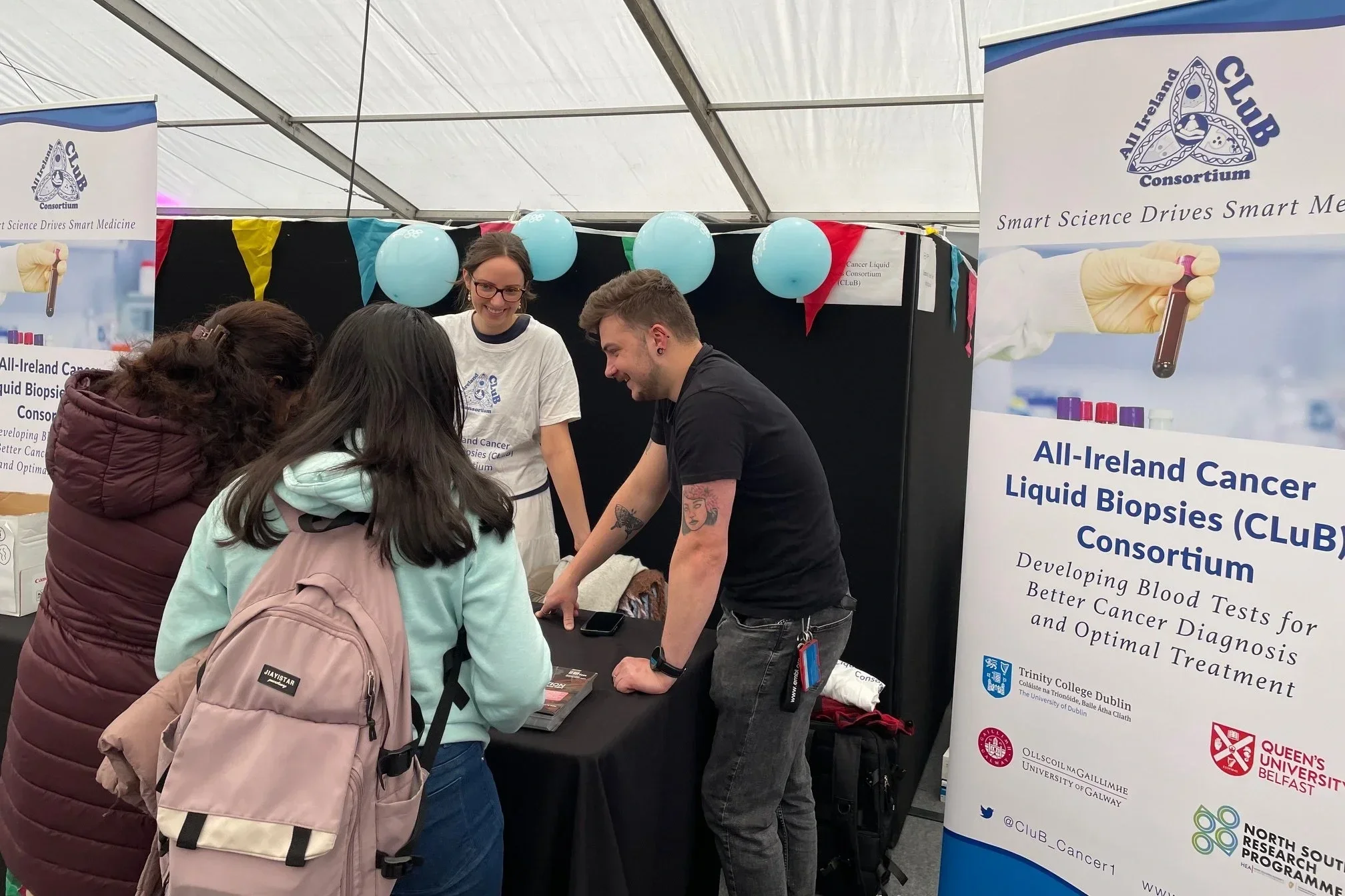People at a booth for the All-Ireland Cancer Liquid Biopsies Consortium, discussing or explaining information related to blood tests for better cancer diagnosis and treatment in a tent with balloons and informational banners.