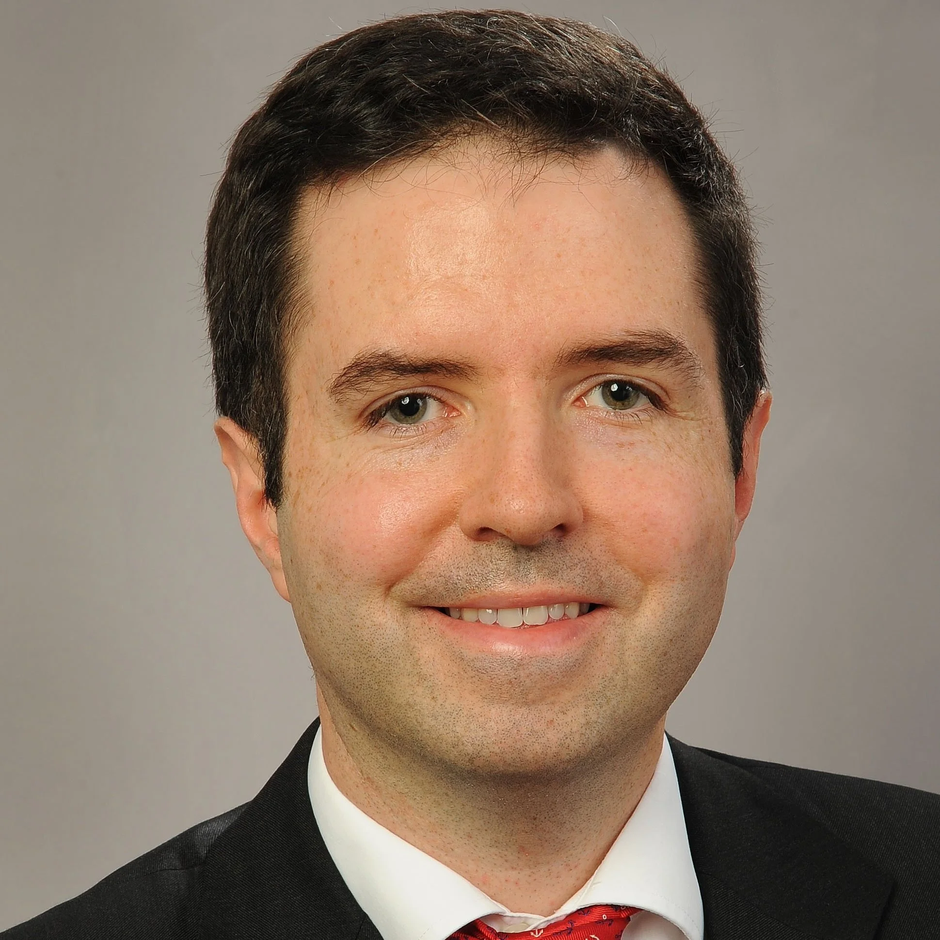 Headshot of a man with dark hair, wearing a black suit, white shirt, and red tie, smiling against a neutral background.