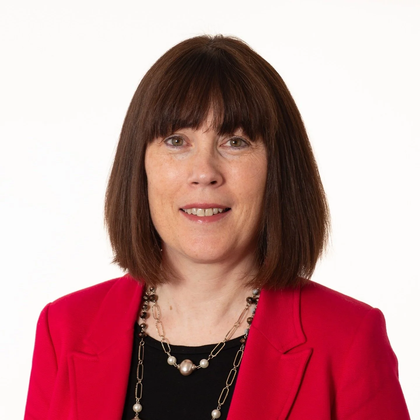A woman with shoulder-length brown hair wearing a red blazer, black top, and a pearl necklace with gold chain accents poses against a white background.
