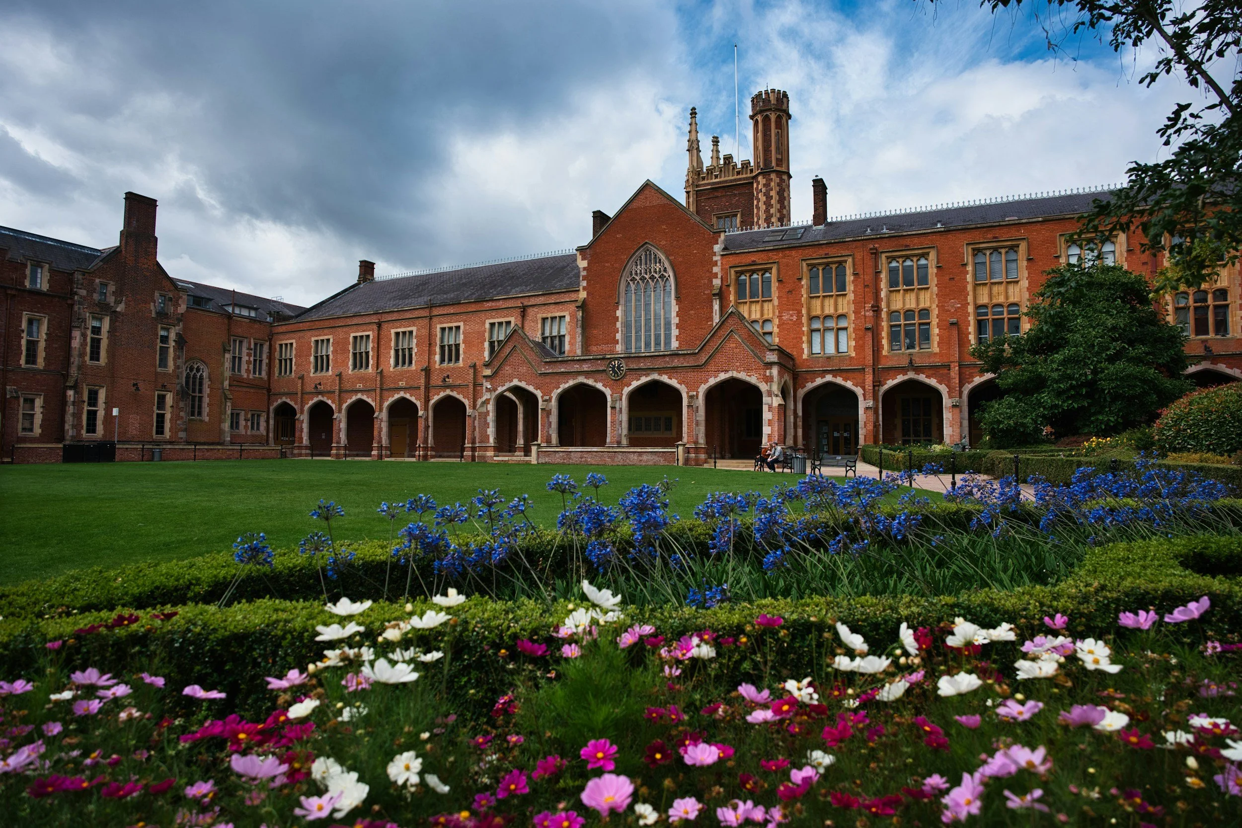 A historic brick building with Gothic windows and arches, set behind a well-maintained lawn and flower beds with pink, white, and purple flowers, beneath a cloudy sky.