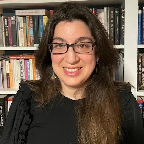 A woman with long brown hair and glasses smiling in front of a bookshelf filled with books.