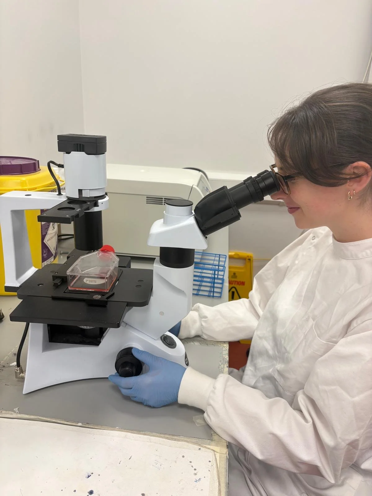 A scientist in safety gloves and glasses looks through a microscope in a laboratory.