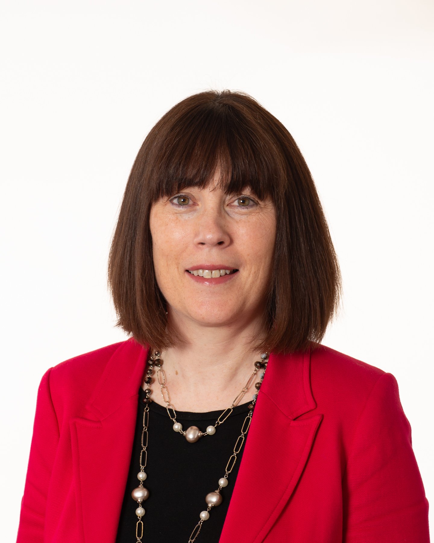Portrait of a woman with shoulder-length brown hair, wearing a red blazer and a pearl necklace, standing against a white background.