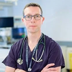 A male doctor wearing glasses and a stethoscope around his neck, standing in a medical office or hospital environment.
