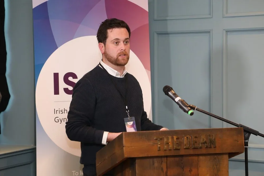 A man with dark hair, beard, wearing a black sweater and white shirt, speaking at a podium with a microphone during a conference or event.