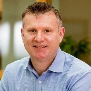 Portrait of a smiling man in a blue shirt with curly hair in an indoor setting.