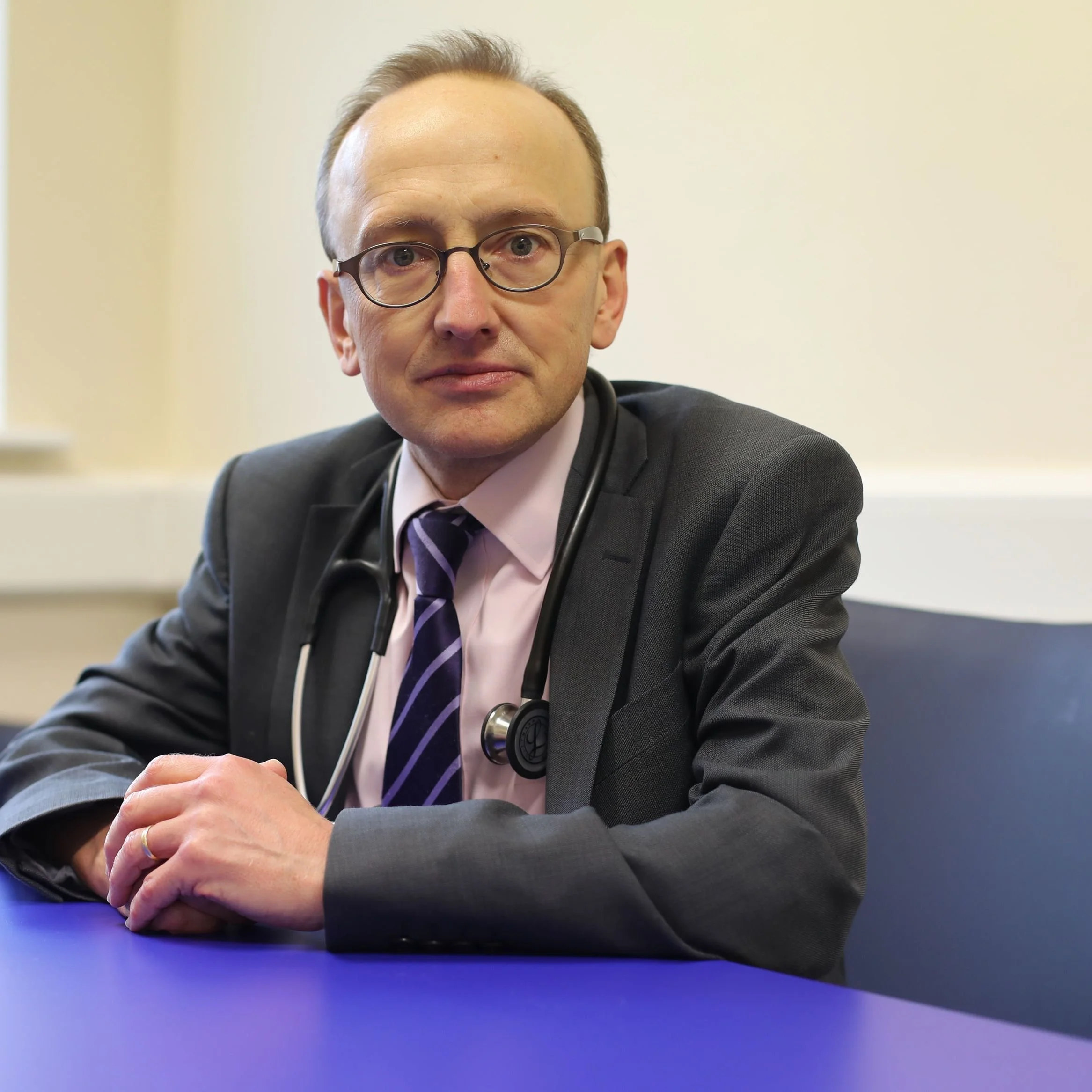 A male doctor or healthcare professional with glasses, dressed in a suit and pink shirt, wearing a stethoscope around his neck, sitting at a blue table in a professional setting, looking at the camera.