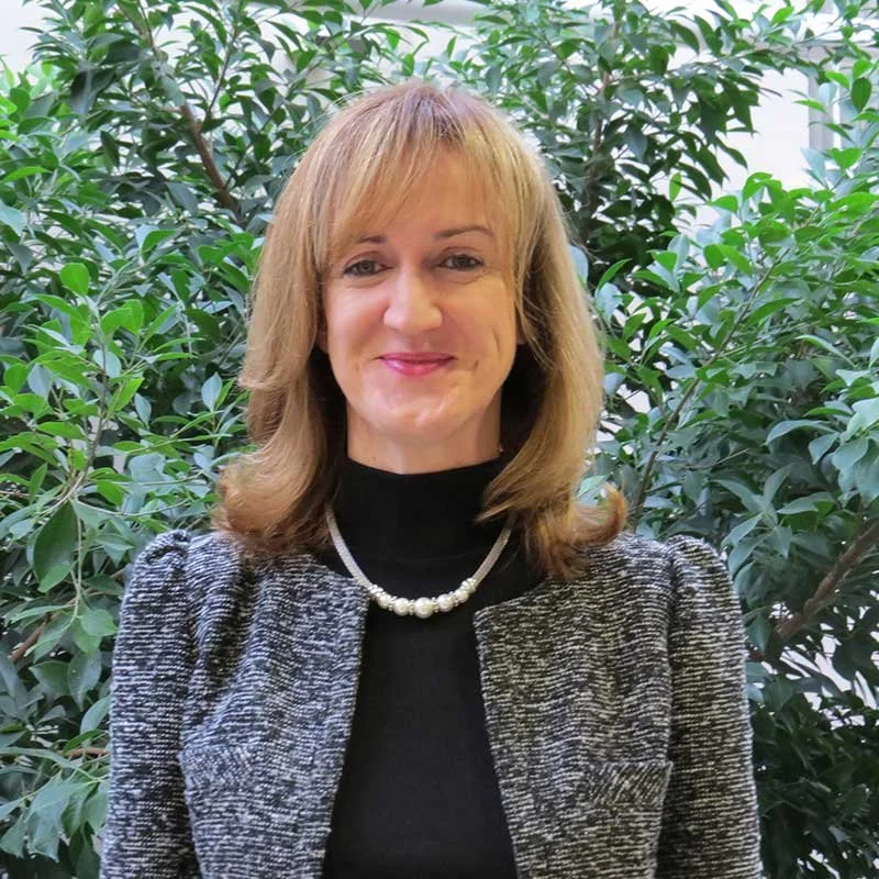 A woman with shoulder-length blonde hair smiling in front of green leafy plants, wearing a black top and a pearl necklace.