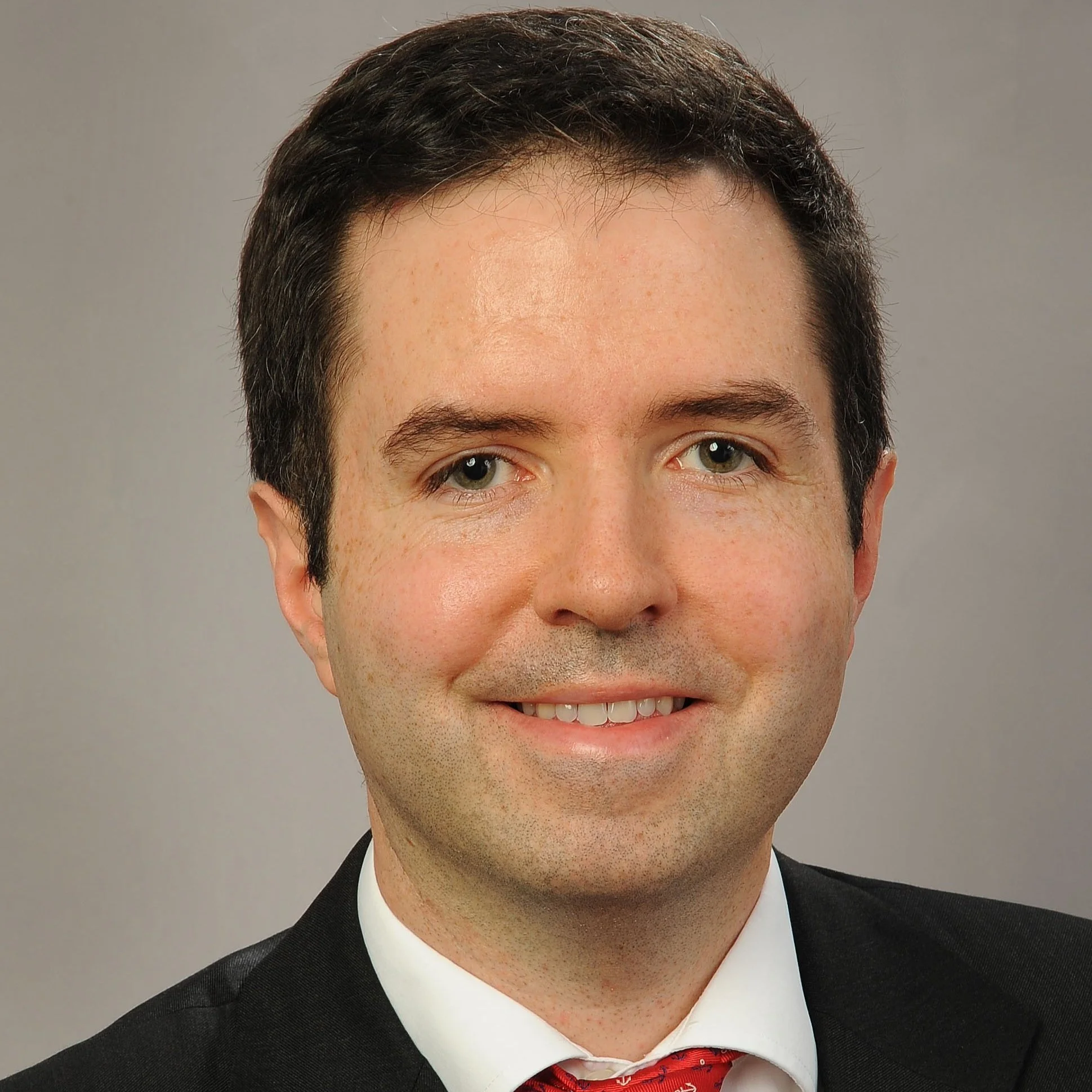 A professional portrait of a man with short dark hair, wearing a black suit, white shirt, and red patterned tie, smiling at the camera against a neutral background.