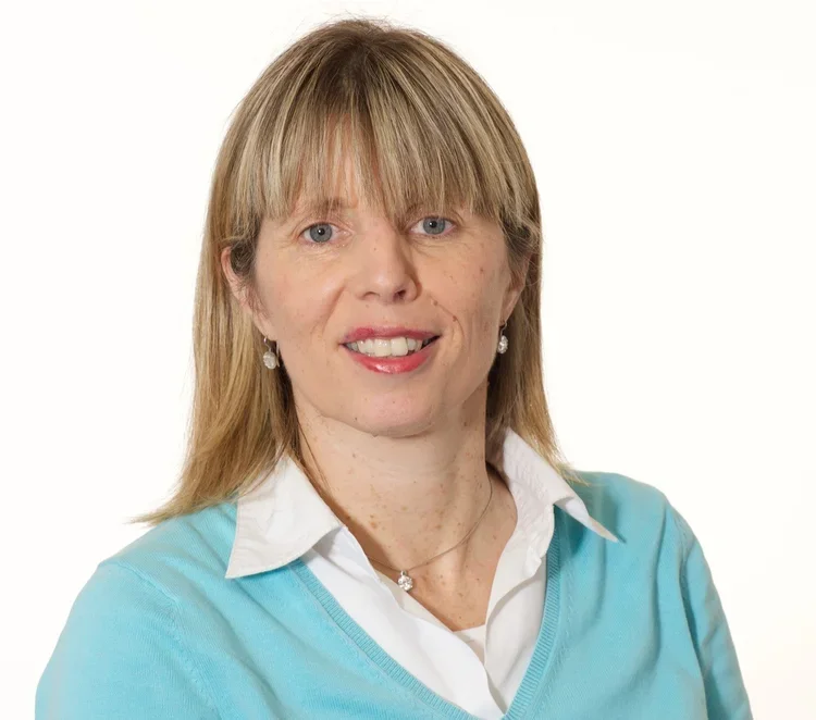A woman with blonde hair and blue eyes smiling, wearing a white collared shirt and a light blue sweater, with earrings and a necklace, against a plain white background.