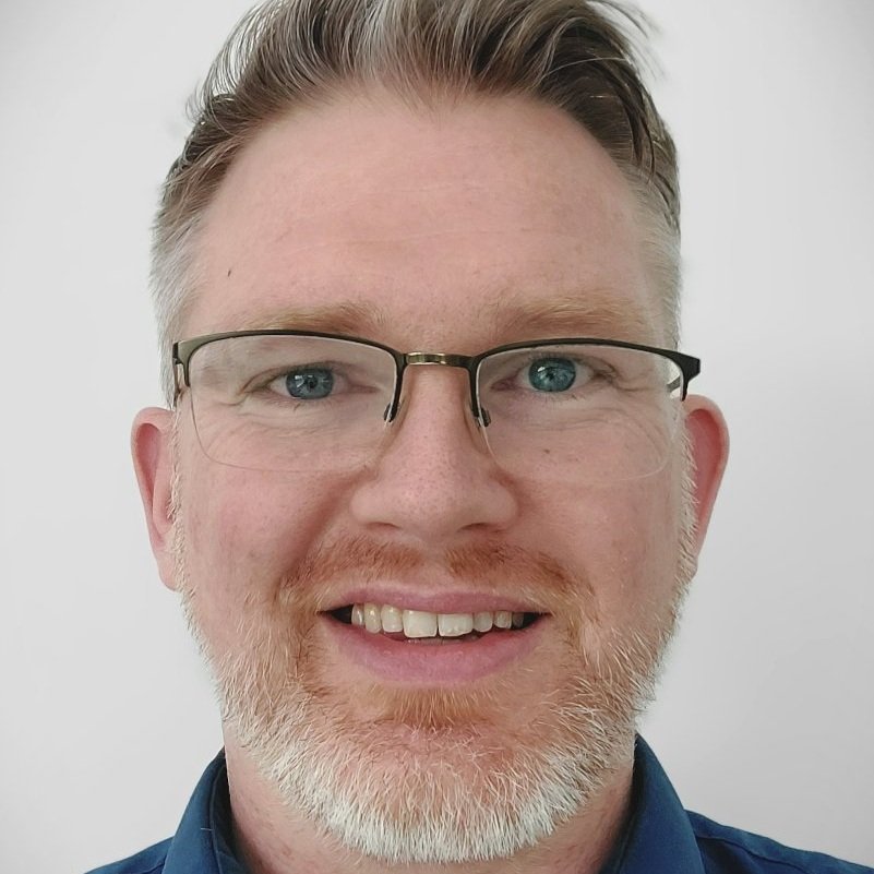 Close-up of a smiling man with glasses, light hair, and a beard against a plain background.
