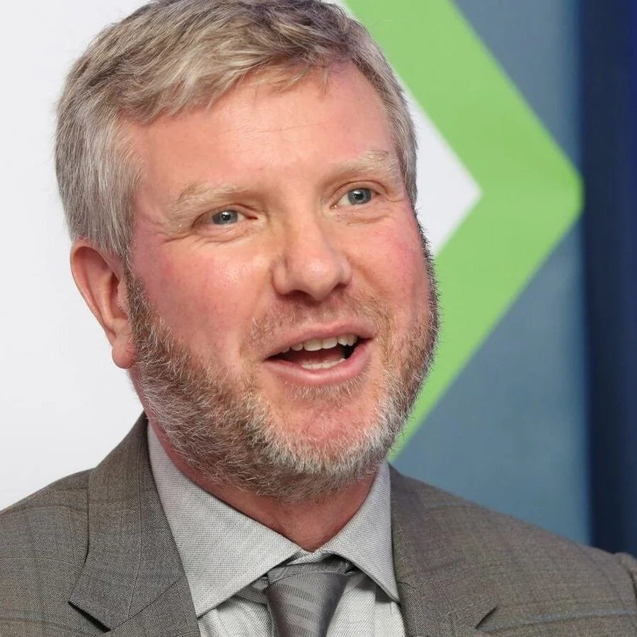 A middle-aged man with gray hair and beard, wearing a gray suit and tie, speaking in front of a background with a green and white geometric design.