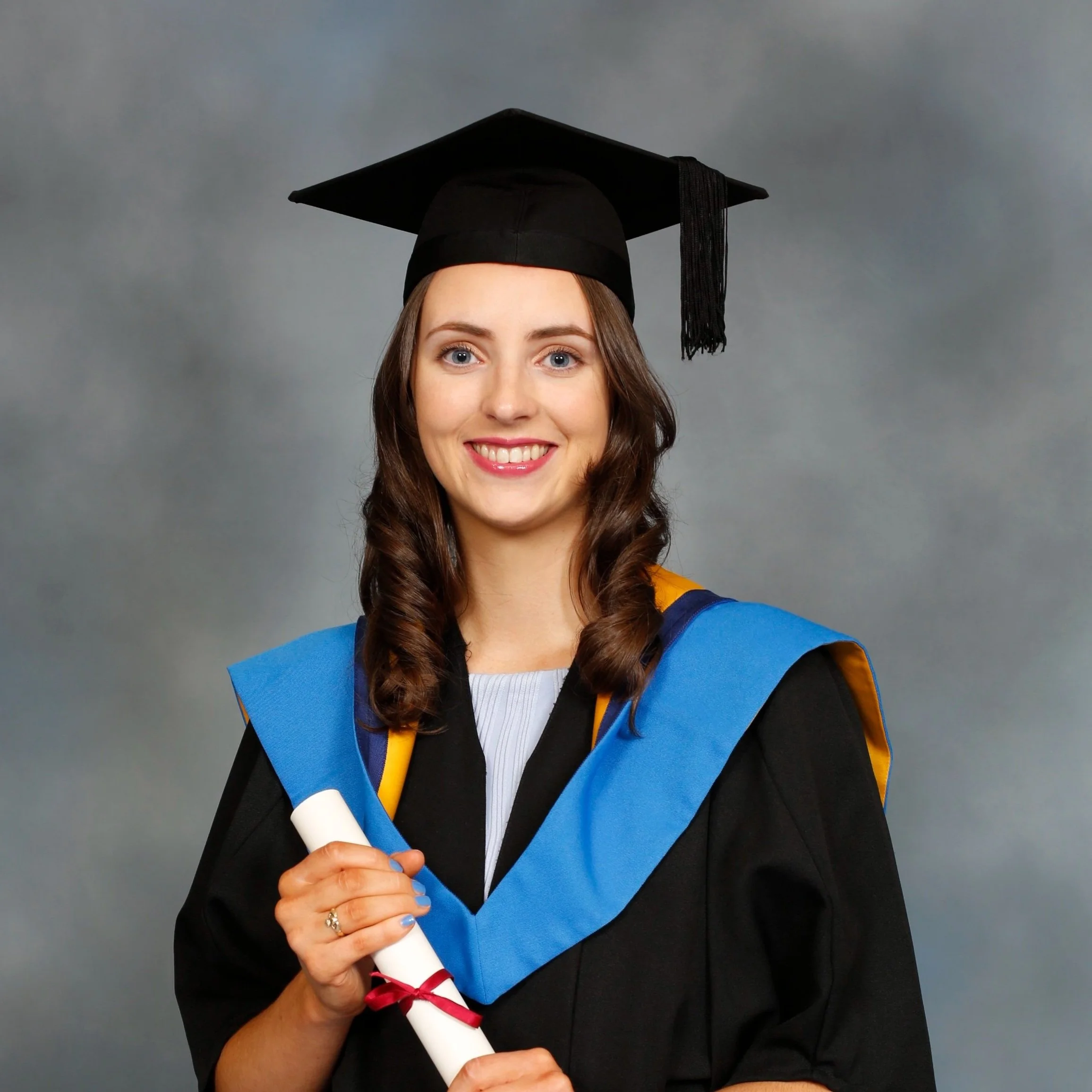 A young woman in a graduation gown wearing a mortarboard hat with a tassel, holding a diploma tied with a red ribbon, smiling confidently against a plain background.