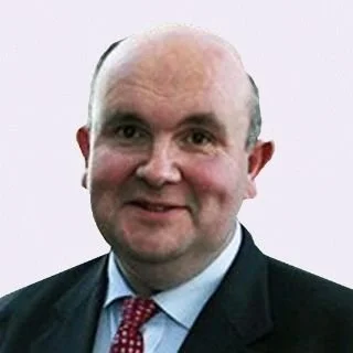 A man with a bald head and a slight smile, dressed in a suit and tie, posing against a plain background.