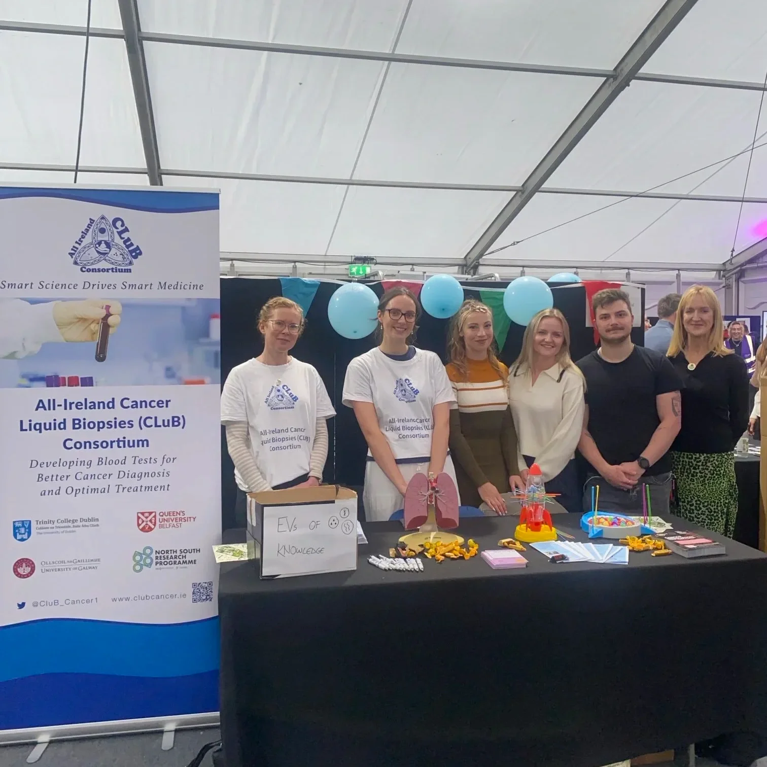A group of six people standing behind a table at an exhibition booth for the All-Ireland Cancer Liquid Biopsies Consortium. The booth has a banner on the left with information about the organization and illustrated scientific themes. The table displays informational materials, a model of lungs, and some small colorful items. The background features balloons and a tent ceiling.