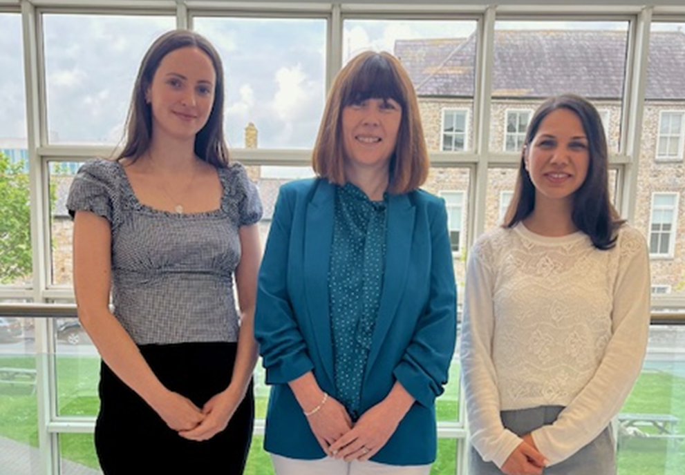 Three women standing inside near large window, with a grassy outdoor area and buildings visible outside.