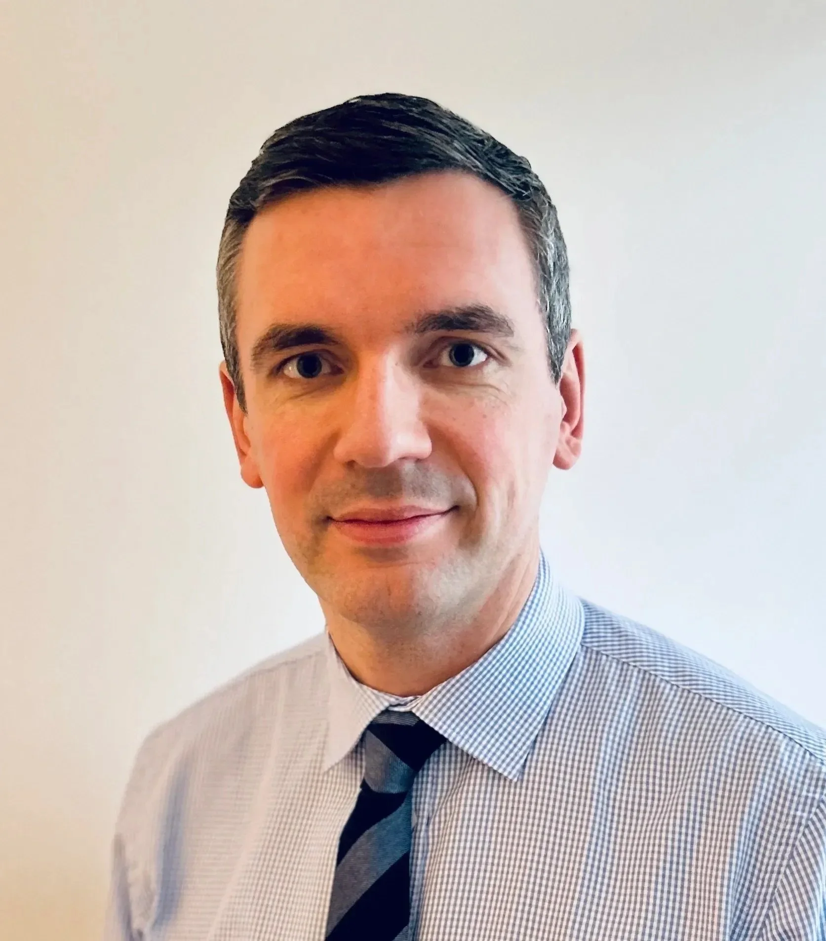 Headshot of a man with short dark hair wearing a light checkered shirt and a dark striped tie against a plain light background.