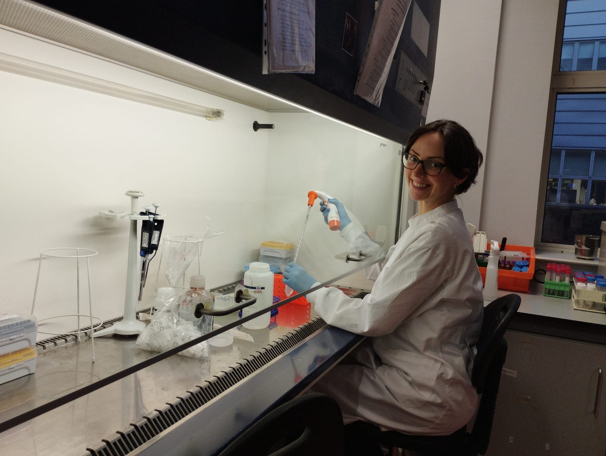 A woman in a white lab coat and glasses working in a laboratory, using a pipette and smiling at the camera.