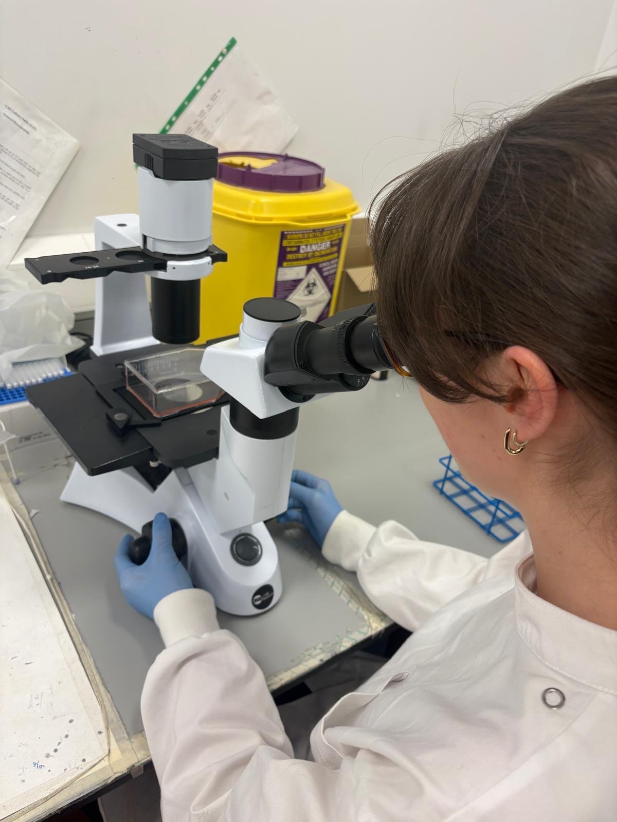A scientist looking through a microscope in a laboratory setting, with lab supplies and safety equipment in the background.