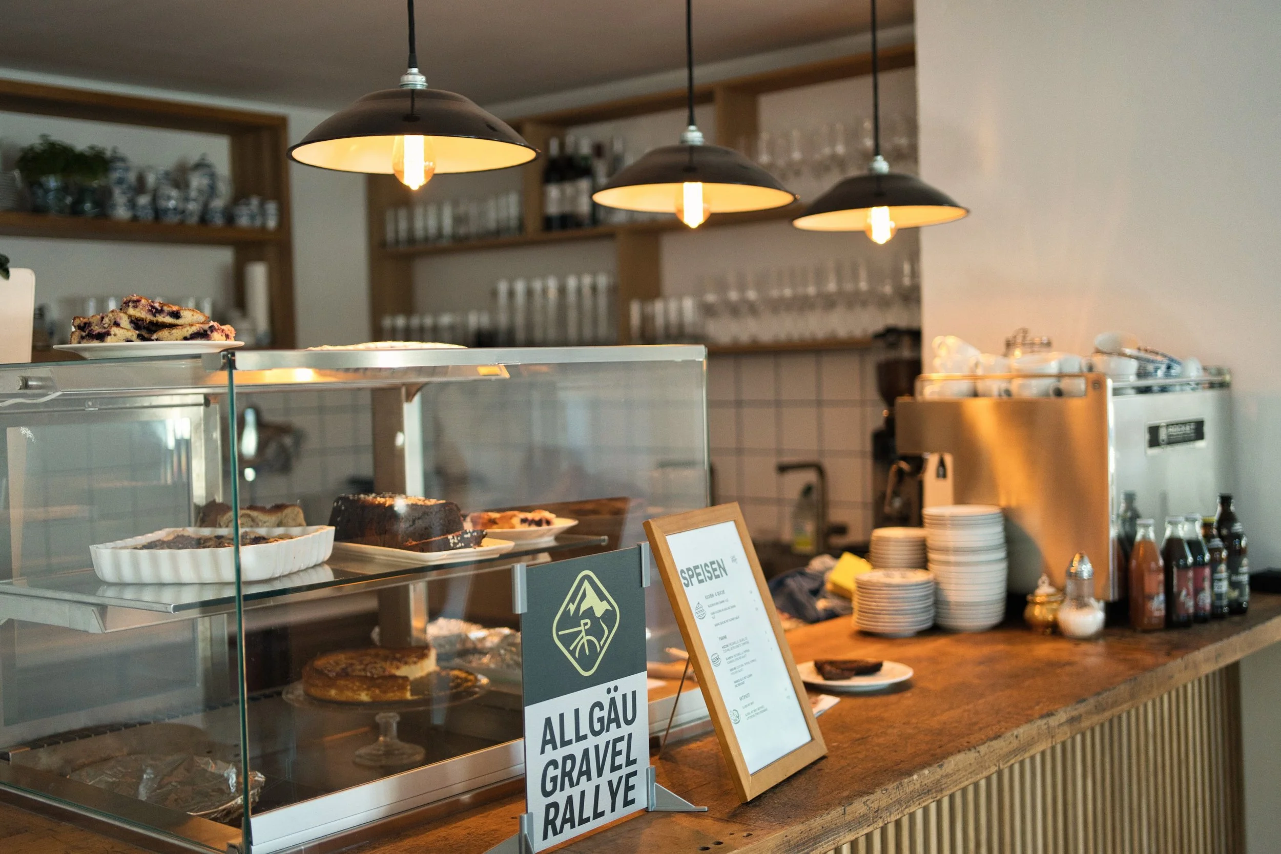 Tresen eines Cafés mit einer Kuchenvitrine , dem Eventschild der Allgäu Gravel Rallye und einer Kaffeemaschine auf der rechten Seite. Über dem Tresen hängen drei schwarze Lampen und im Hintergrund sind Gläser in einem Regal zu sehen.