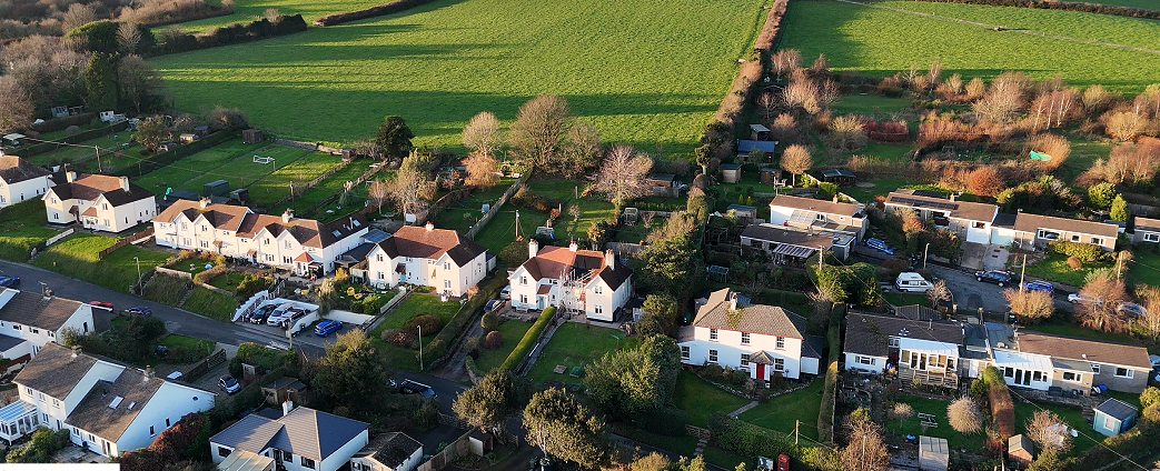Aerial view of a neighborhood with houses, gardens, and trees, surrounded by green fields and farmland.