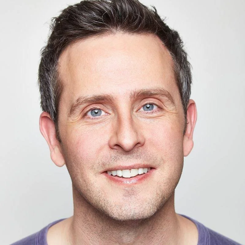Headshot of a smiling man with blue eyes and dark brown hair, wearing a purple shirt, against a white background.