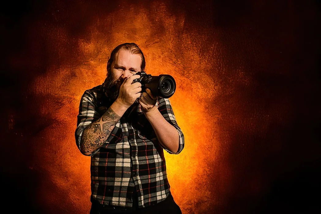 A man with a beard and tattoos on his arm is holding a camera and taking a photo. He is wearing a plaid shirt and is standing against a fiery orange and dark background.