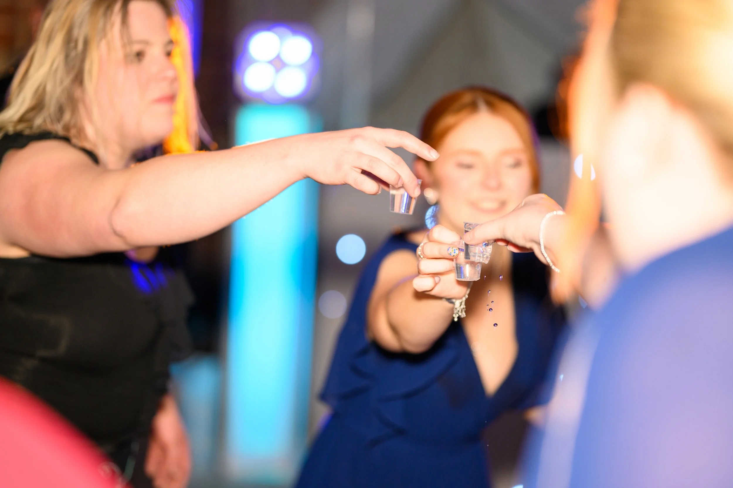 Three women at a party are pouring clear liquid into shot glasses. The woman on the left is wearing a black top, the woman in the middle has red hair and is smiling, and the woman on the right, seen partially, is wearing a blue dress. The background has colorful lights and a blurry atmosphere.