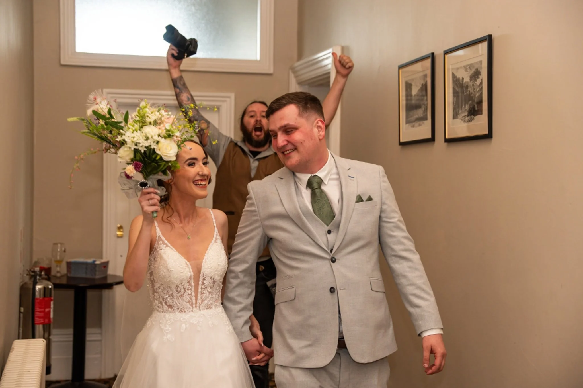 A bride and groom holding hands and smiling, walking down a hall during a wedding celebration, with a person behind them cheering and raising a camera.