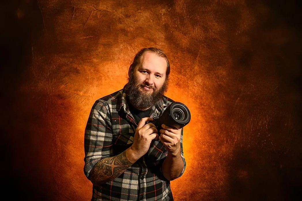 A man with a beard and tattoos, wearing a plaid shirt, holding a professional camera with a large lens, posing against a warm, textured brown background.