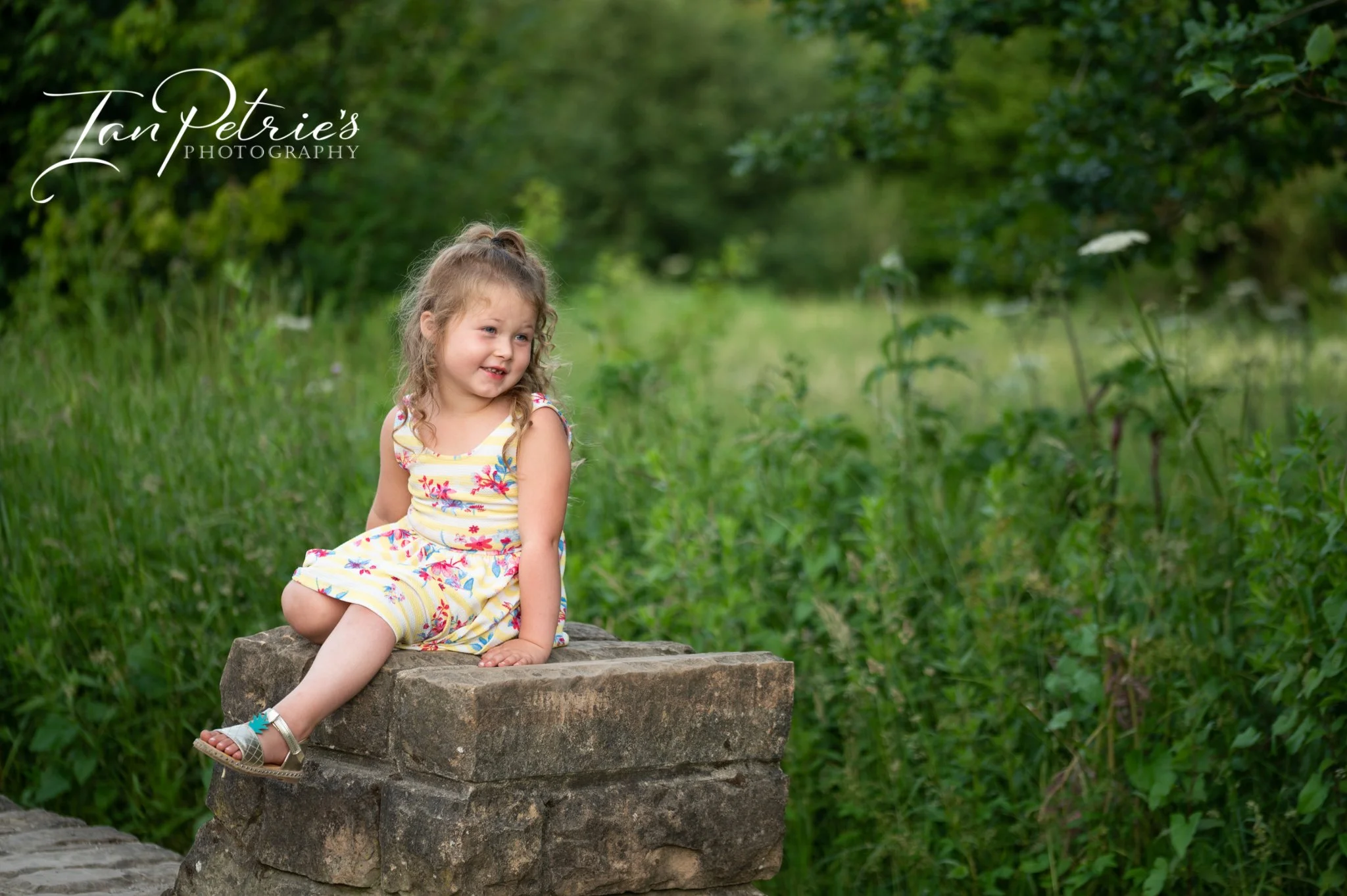 A young girl with curly hair in a colorful striped dress sitting on a rock by a stream in a lush green natural setting.