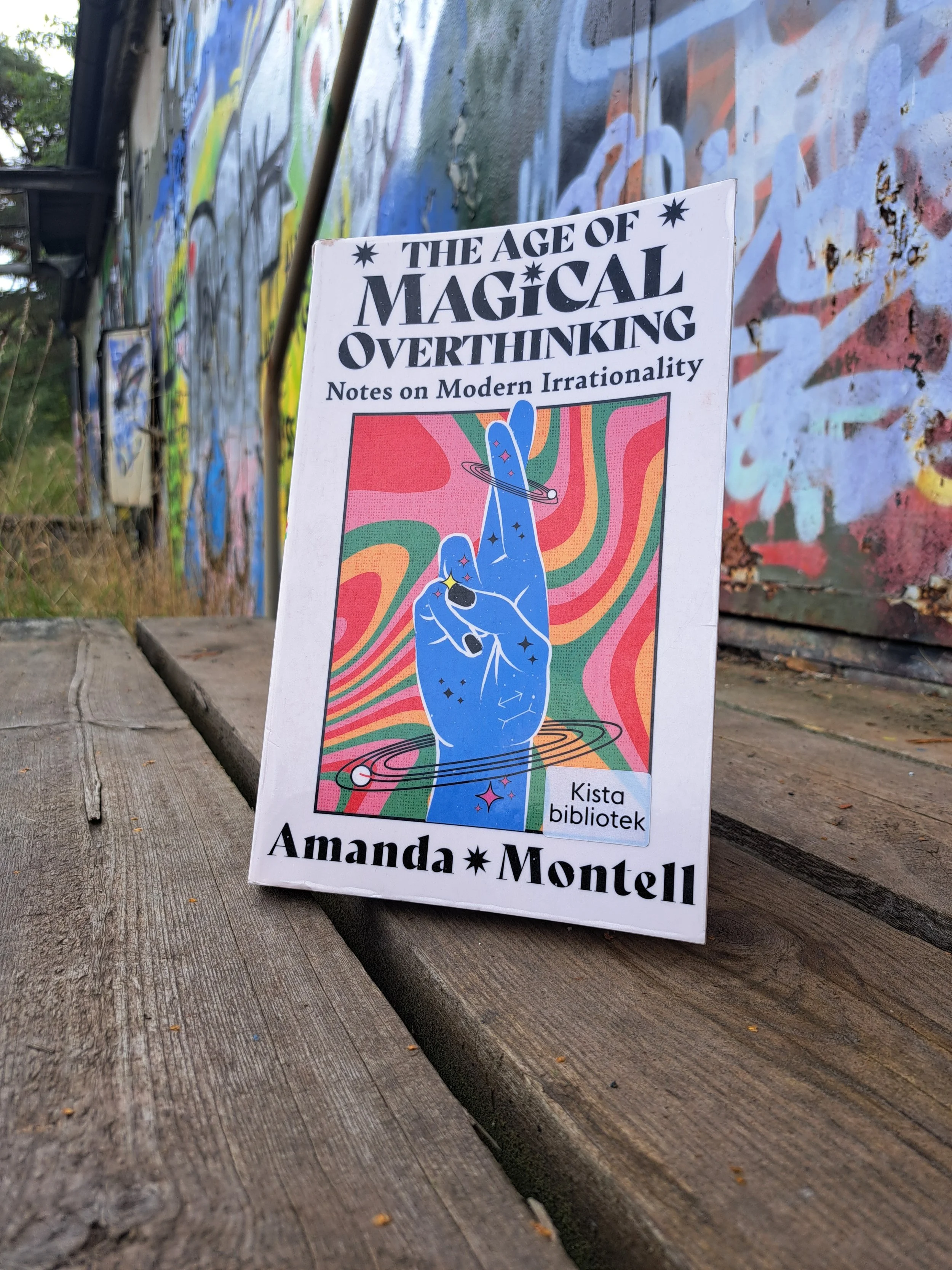 The book The Age Of Magical Overthinking by Amanda Montell sat on a wooden floor by a heavily graffitied wall.