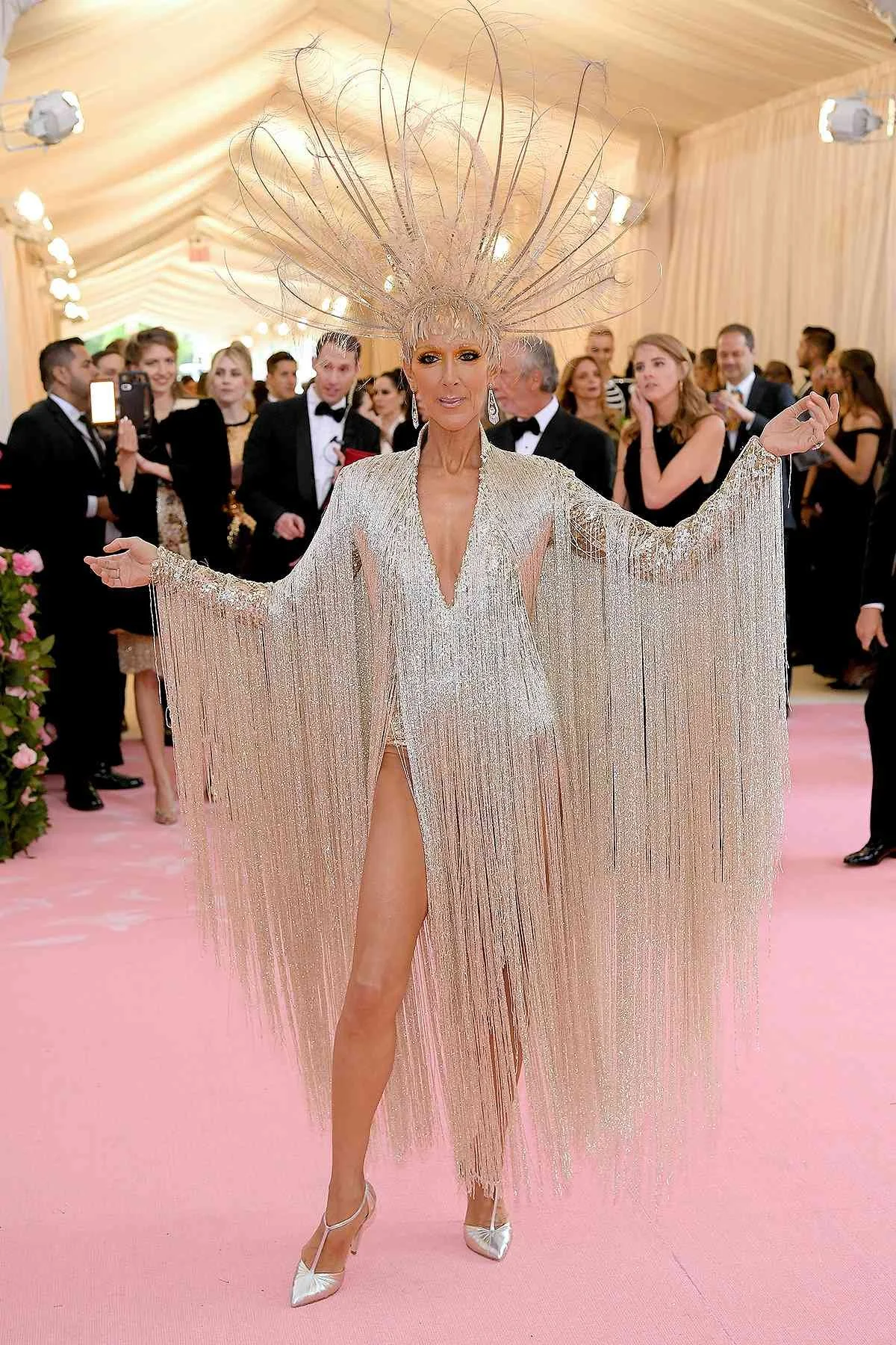 Celine Dion wearing a very sparkly dress with a massive feather headpiece both in silver, in front of a crowd of people dressed in black